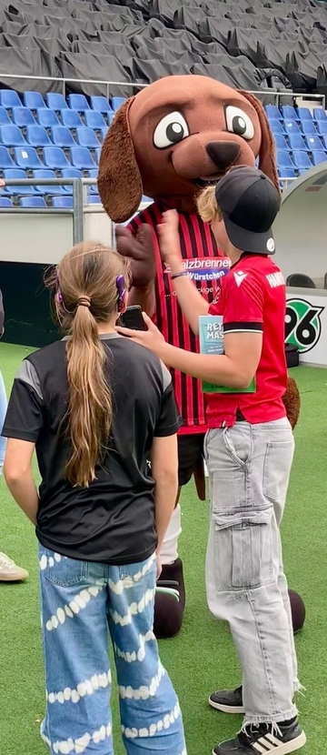 Bild zeigt das Maskottchen EDDI mit einer Schülerin und einem Schüler im Stadion