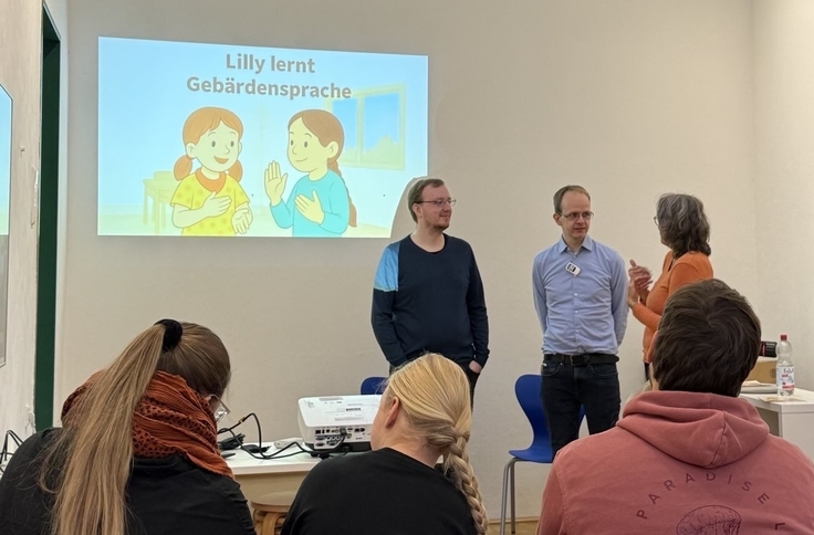 Ein Bildschirm zeigt den Titel eines Buches: Lilly lernt Gebärdensprache. Die vortragende Personengruppe aus einer tauben Person, dem Autor des Buches und einer dolmetschende Person. Im Vordergrund sind drei Zuhörende abgebildet.