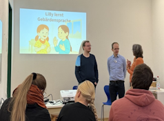 Ein Bildschirm zeigt den Titel eines Buches: Lilly lernt Gebärdensprache. Die vortragende Personengruppe aus einer tauben Person, dem Autor des Buches und einer dolmetschende Person. Im Vordergrund sind drei Zuhörende abgebildet.