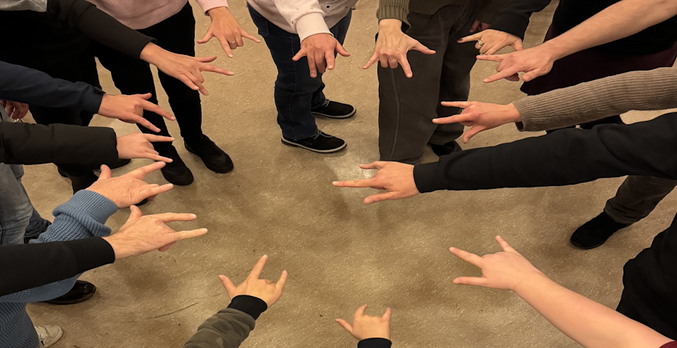 Auf dem Bild sind 15 zur Mitte gesteckte Hände abgebildet, die die Gebärde I-love-You gebärden.