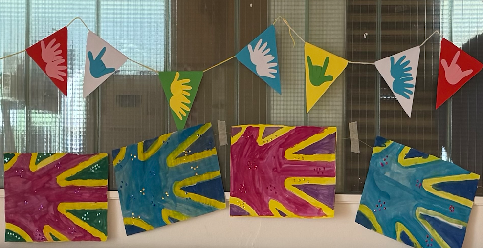 Auf dem Bild sieht man die Deaf Flag von Kindern in verschiedenen Farben gestaltet. Über den verschiedenen Deaf Flags hängen noch Wimpel mit gebärdenden Händen.